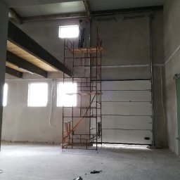 Tomasz Piecyk voltom instalacje elektryczne pomiary - Montaż bramy garażowej segmentowej: monter na rusztowaniu przytwierdza prowadnicę do ściany. W tle widać okna i surowy stan wykończenia wnętrza.