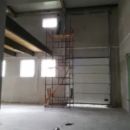 Montaż bramy garażowej segmentowej: monter na rusztowaniu przytwierdza prowadnicę do ściany. W tle widać okna i surowy stan wykończenia wnętrza.