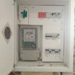 Otwarta skrzynka z licznikiem i bezpiecznikami wewnątrz, widoczne przewody. Biała obudowa na tle ściany w trakcie remontu. Widok z bliska na instalację elektryczną.