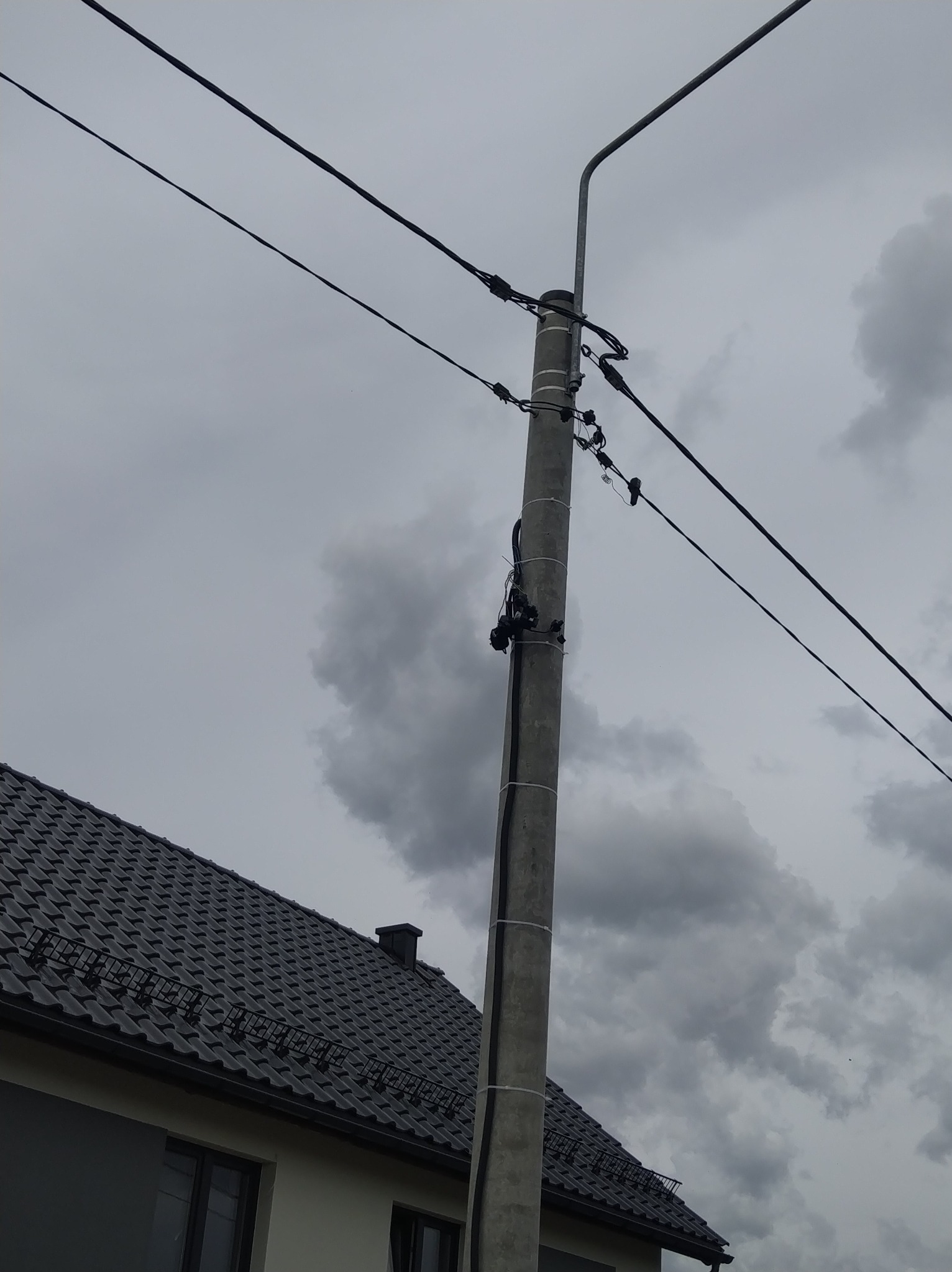 Słup elektryczny z przyłączem kablowym na tle pochmurnego nieba w Szabdzie. Widoczny fragment dachu domu z ciemną dachówką.