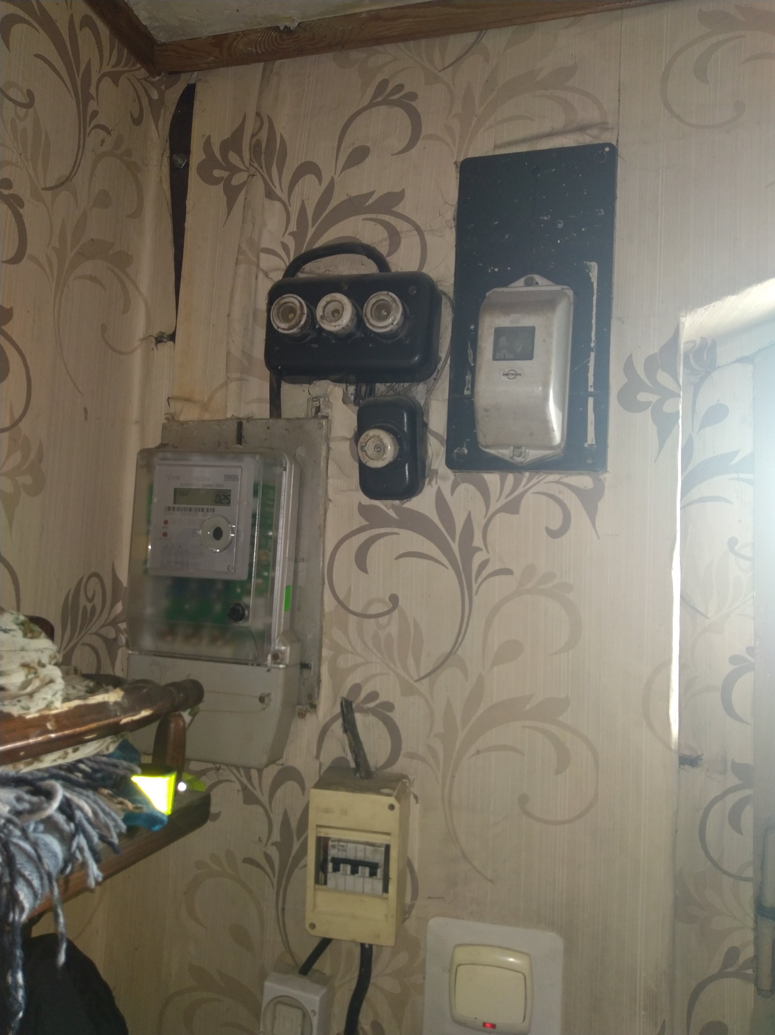 Stara instalacja elektryczna na ścianie z wzorzystą tapetą. Widoczne: licznik, bezpieczniki topikowe, wyłącznik różnicowoprądowy i włącznik światła. Niefachowy montaż.