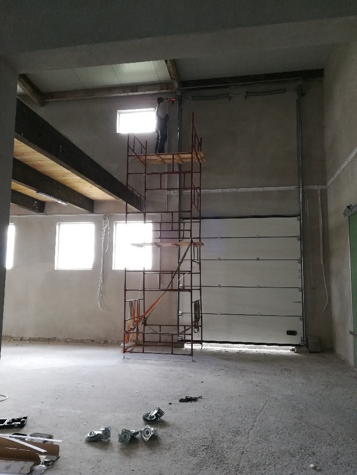 Montaż bramy garażowej segmentowej: monter na rusztowaniu przytwierdza prowadnicę do ściany. W tle widać okna i surowy stan wykończenia wnętrza.