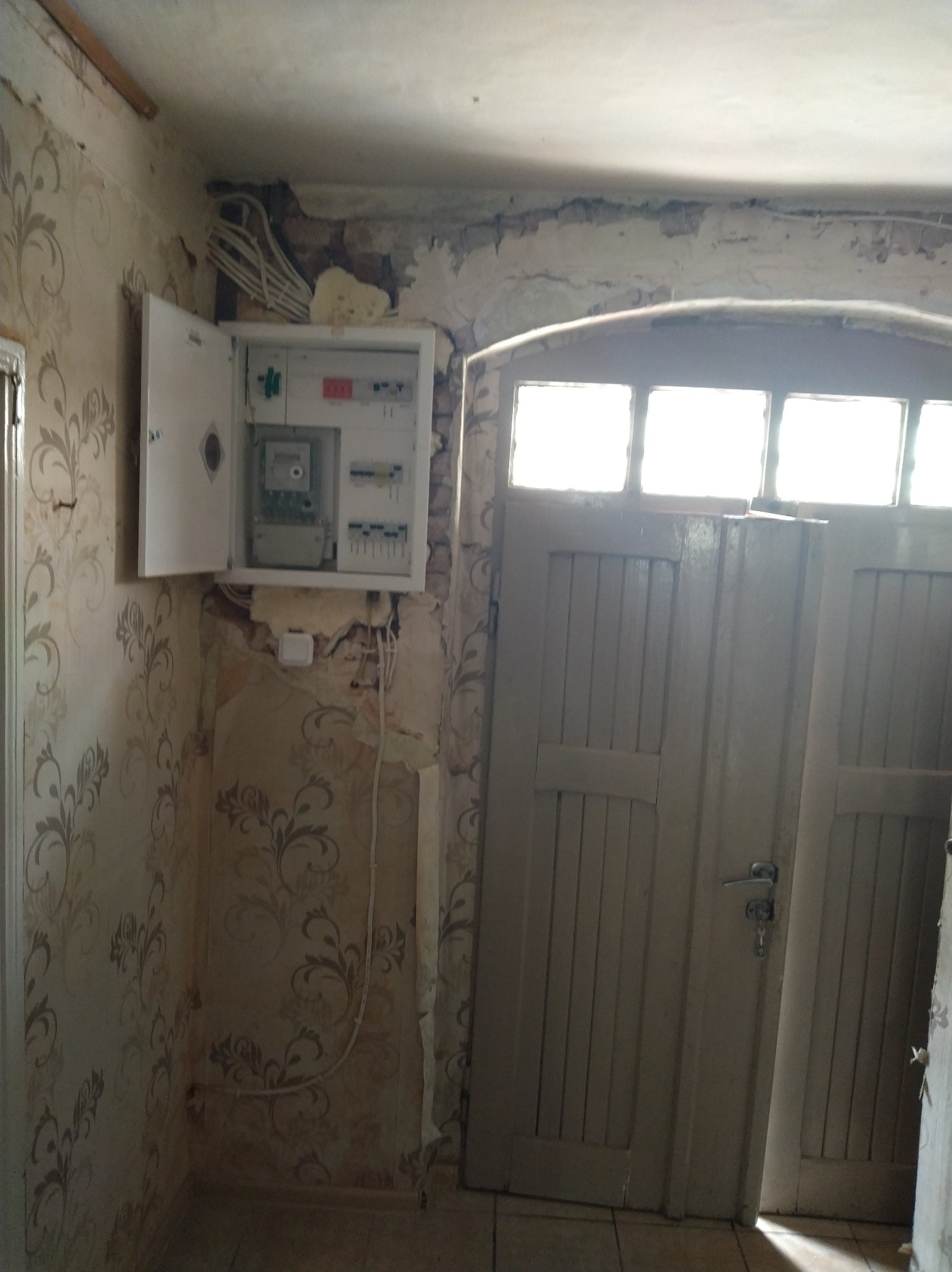 Otwarta skrzynka z licznikiem i bezpiecznikami na ścianie z tapetą, obok drewniane drzwi z szybami. Widoczne kable i fragmenty instalacji elektrycznej.