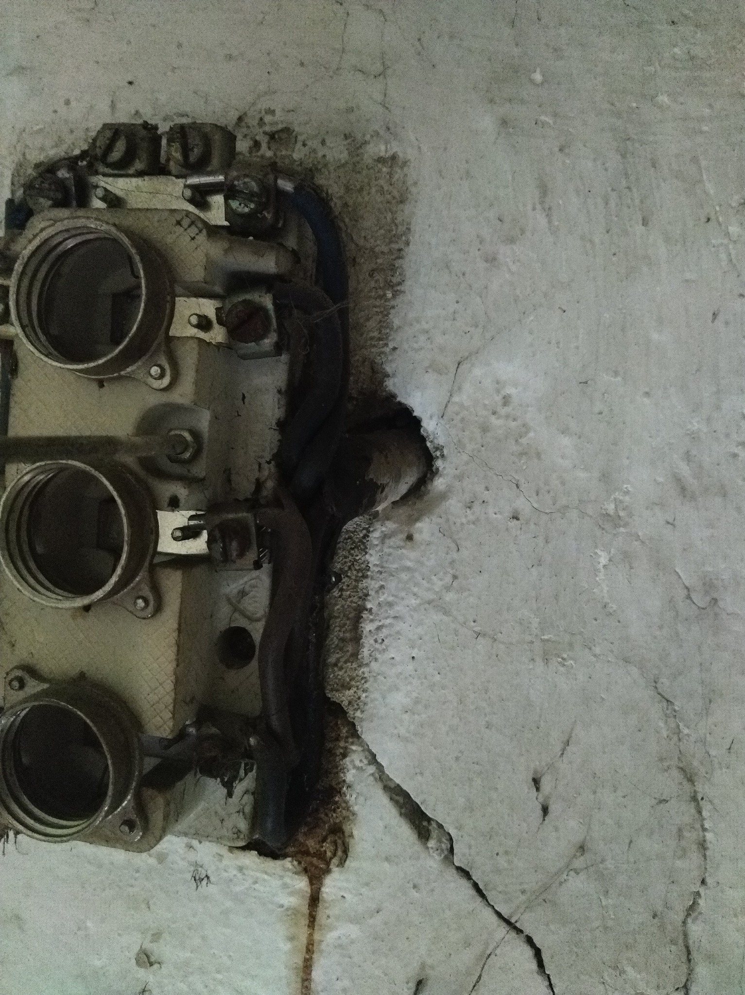 Stara, uszkodzona skrzynka z bezpiecznikami, wtopiona w popękany, szary tynk. Widoczne przewody i elementy montażowe. Niefachowe wykończenie, ślady korozji.