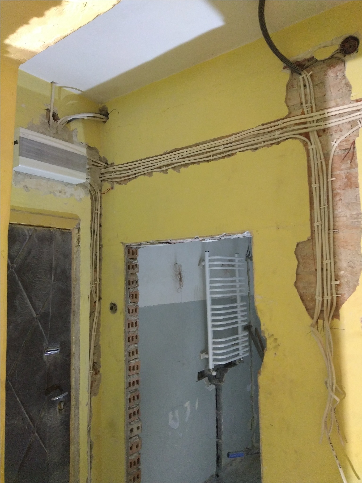 Żółta ściana w trakcie remontu z widocznymi przewodami elektrycznymi ułożonymi w bruzdach i wystającą cegłą, grzejnik w tle.