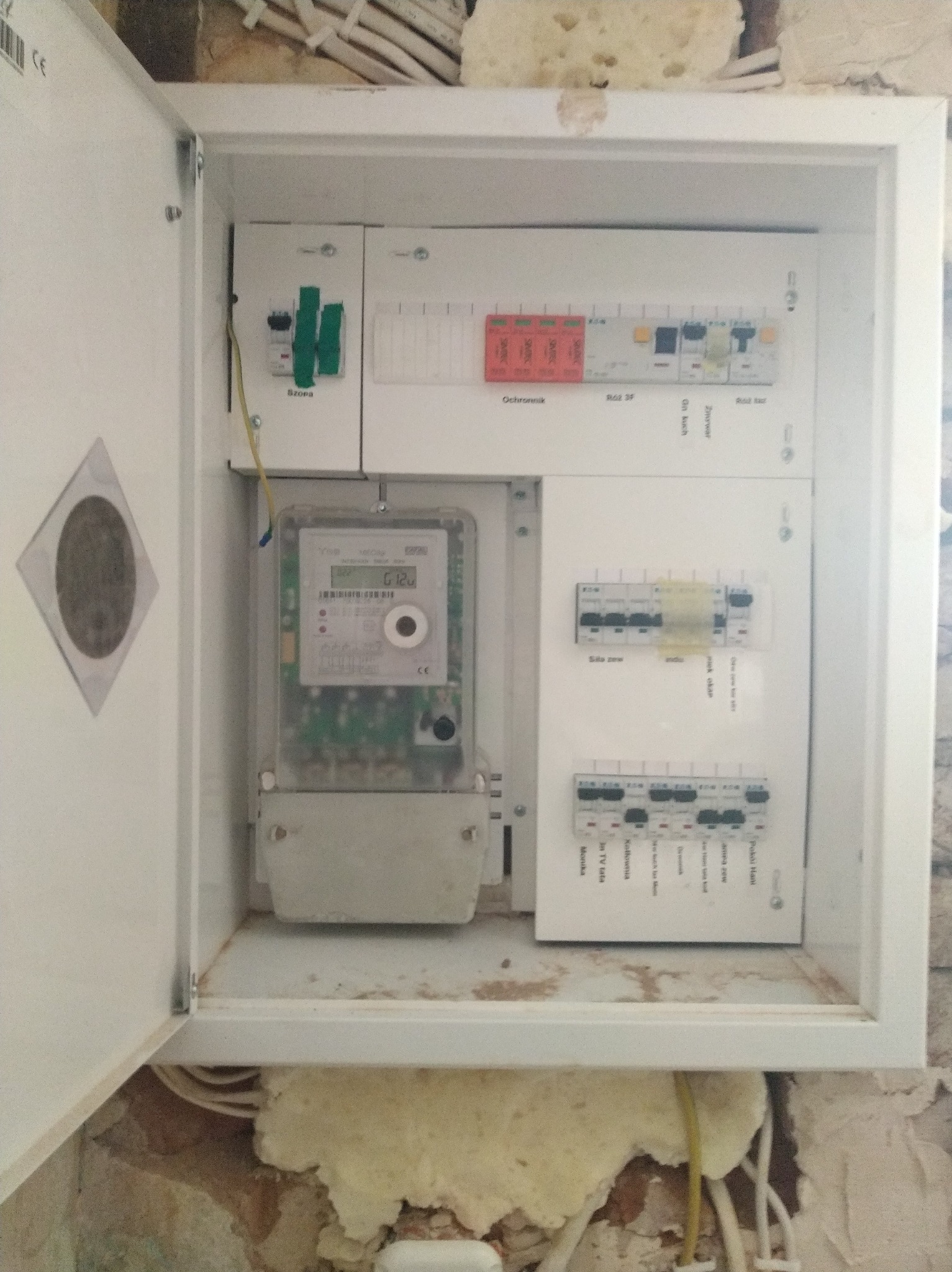 Otwarta skrzynka z licznikiem i bezpiecznikami wewnątrz, widoczne przewody. Biała obudowa na tle ściany w trakcie remontu. Widok z bliska na instalację elektryczną.