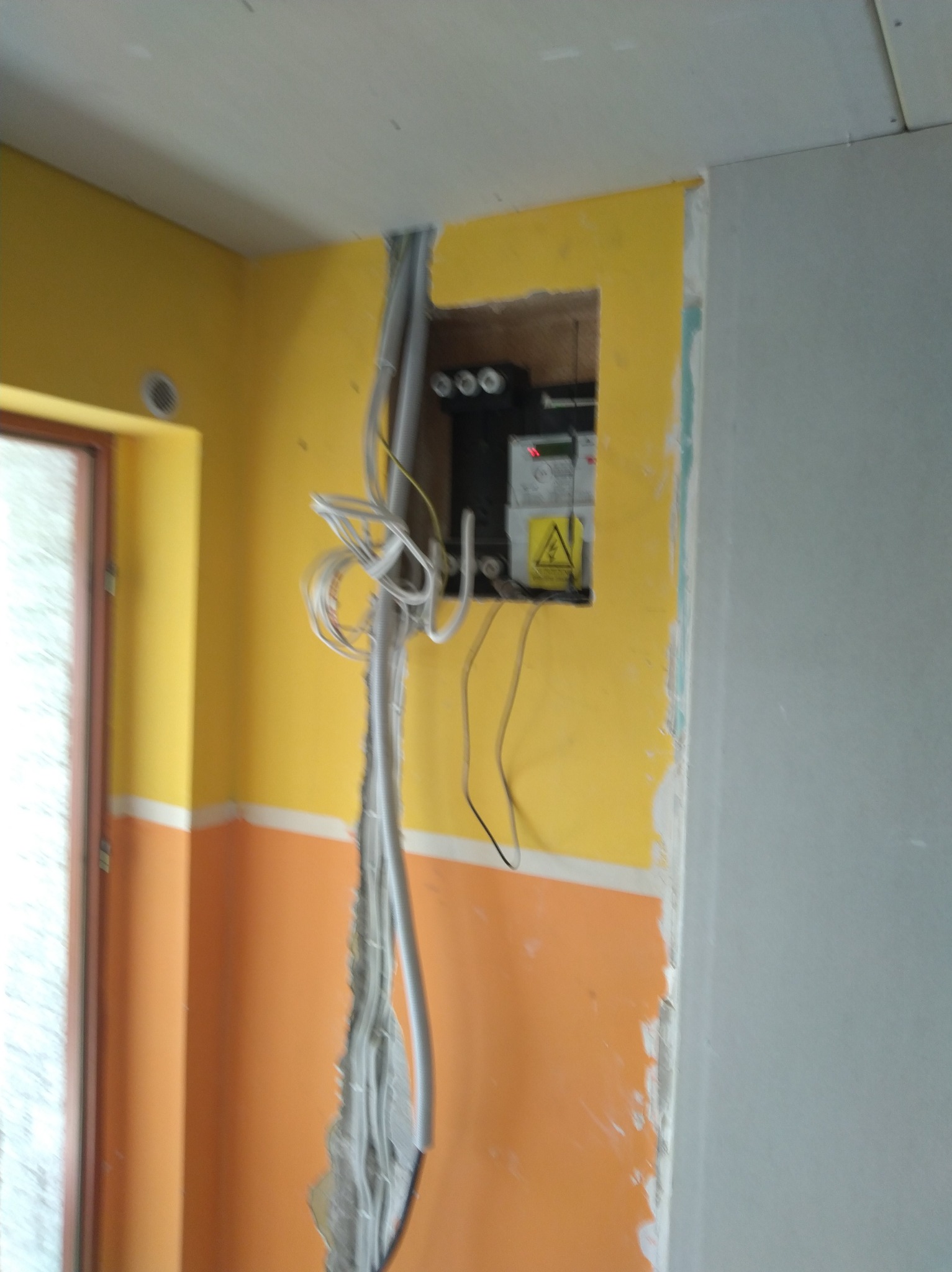 Niedokończona instalacja elektryczna w ścianie z widoczną puszką i kablami na żółto-pomarańczowym tle, obok okno i szara ściana w trakcie prac wykończeniowych.