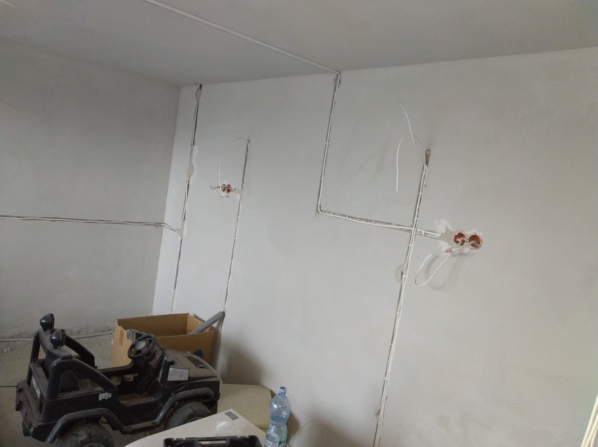 Białe kable elektryczne prowadzone po ścianie w nowym budynku. Widoczne puszki instalacyjne. W tle zabawkowy samochód i karton.
