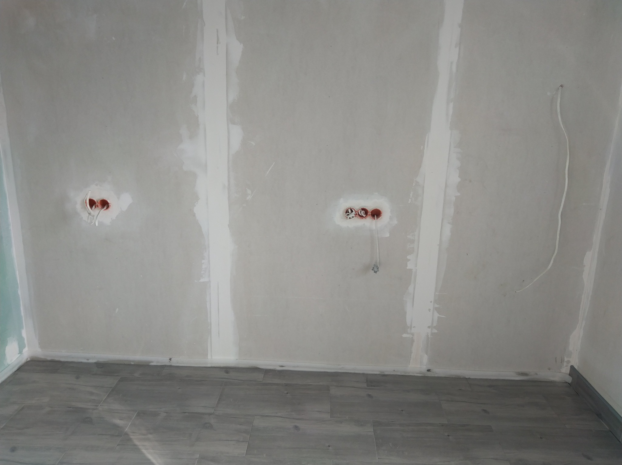 Ściana w trakcie prac wykończeniowych z widocznymi puszkami instalacyjnymi i wystającymi przewodami w nowym pomieszczeniu z szarą podłogą.