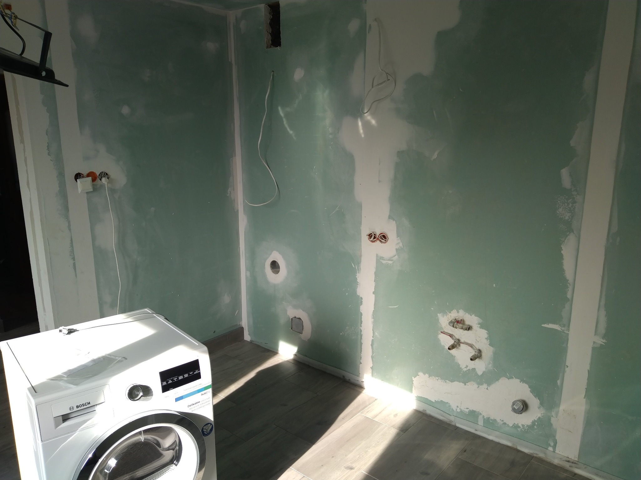 Wnętrze w trakcie remontu: ściany z płyt gipsowych, instalacja elektryczna i hydrauliczna przygotowana pod montaż pralki Bosch, widoczne przewody i gniazdka.