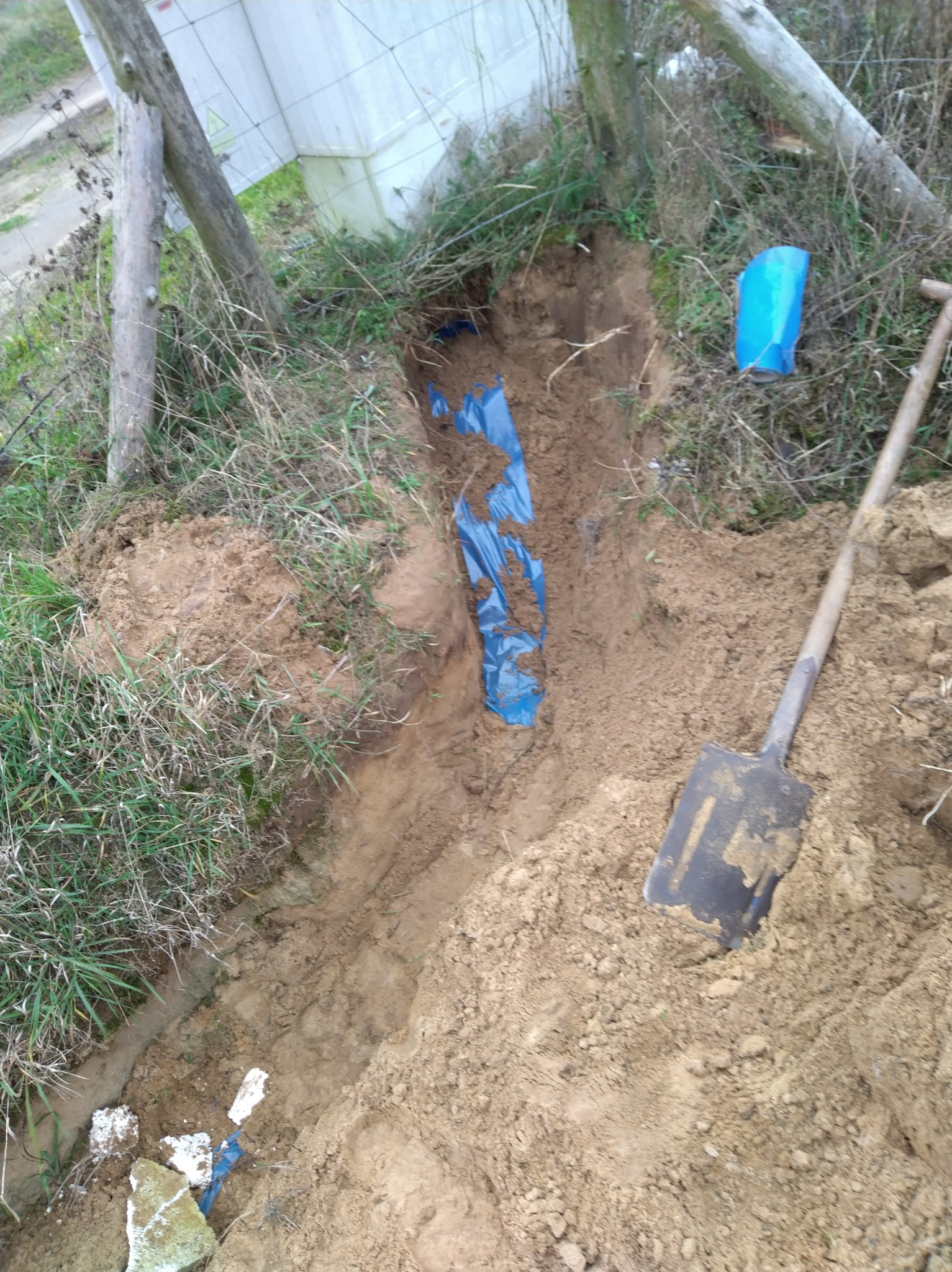 Wykop w ziemi z niebieską folią zabezpieczającą, widoczna łopata i fragment białej konstrukcji. Prace ziemne związane z instalacją podziemną.