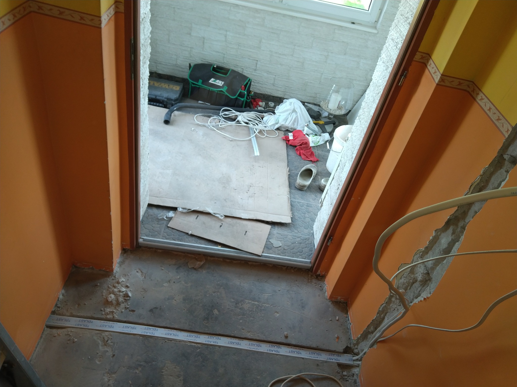 Remont korytarza: widok z góry na rozłożone kable, narzędzia i materiały budowlane przy wejściu do pomieszczenia z oknem. Pomarańczowe ściany w trakcie prac.