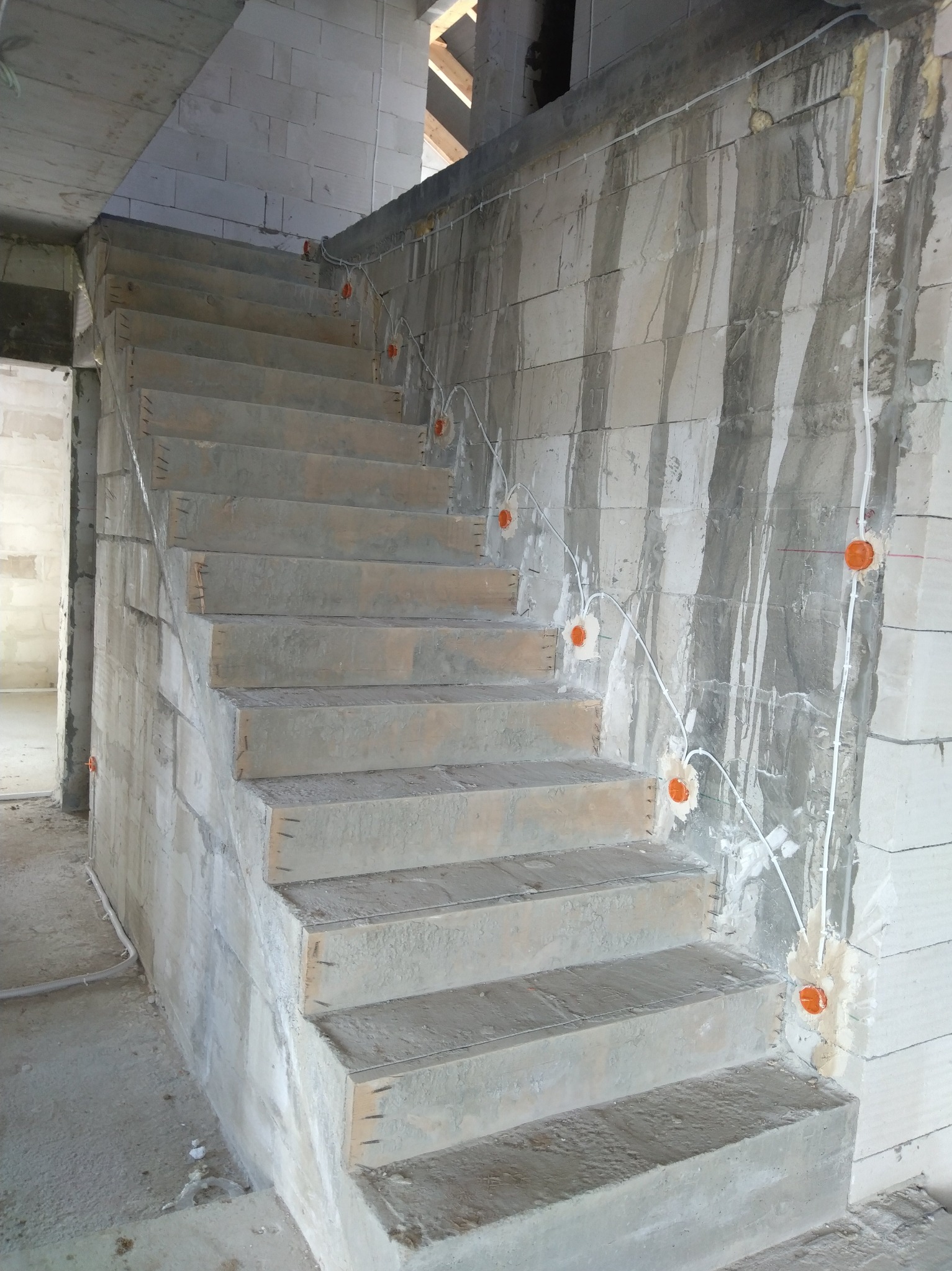 Surowe schody betonowe w trakcie budowy z widoczną instalacją elektryczną w ścianie. Pomarańczowe puszki elektryczne rozmieszczone wzdłuż biegu schodów.