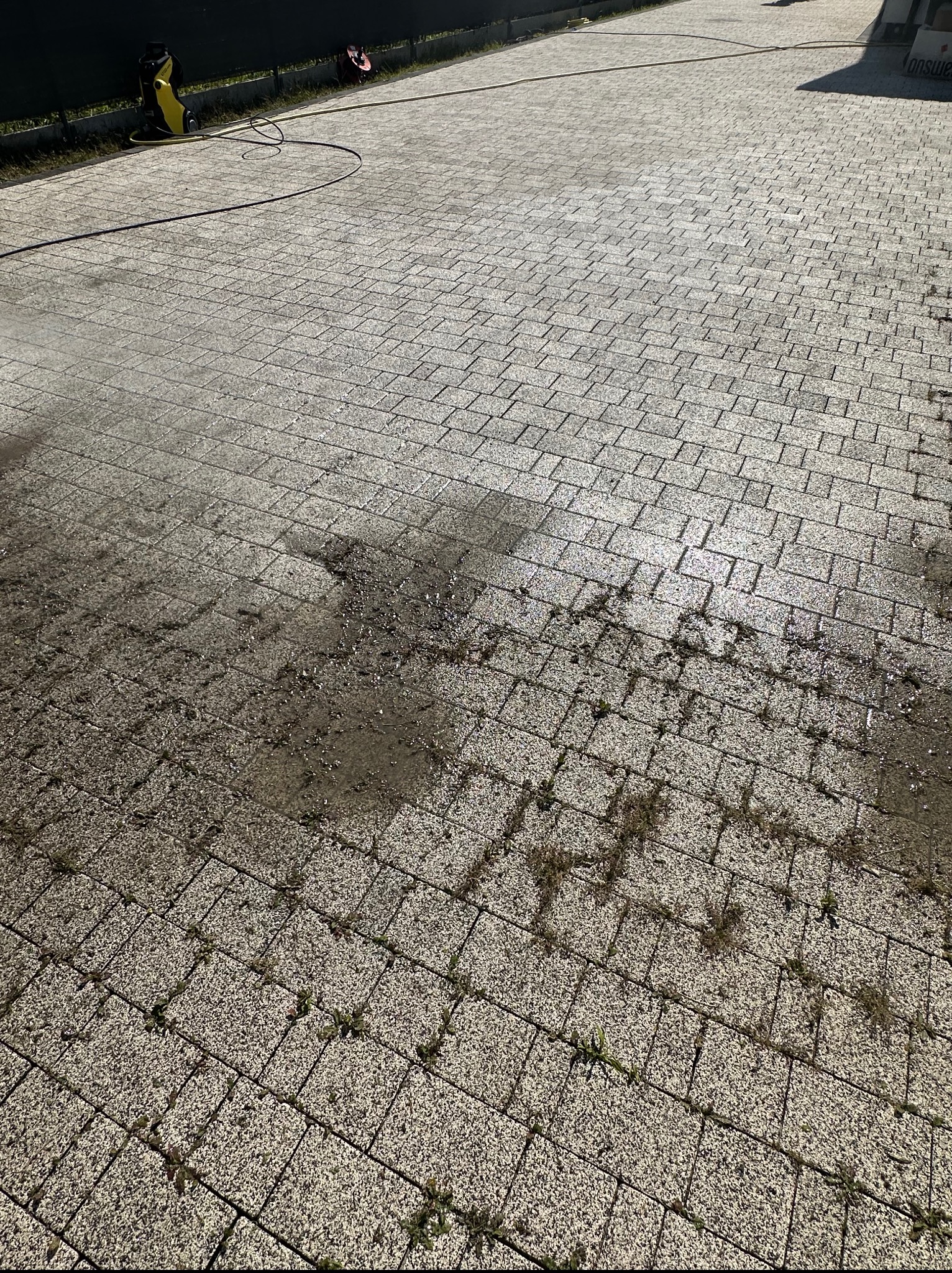 Częściowo umyta kostka brukowa z resztkami brudu i roślinności, widoczny sprzęt do mycia ciśnieniowego w tle. Kontrast czystej i brudnej powierzchni.