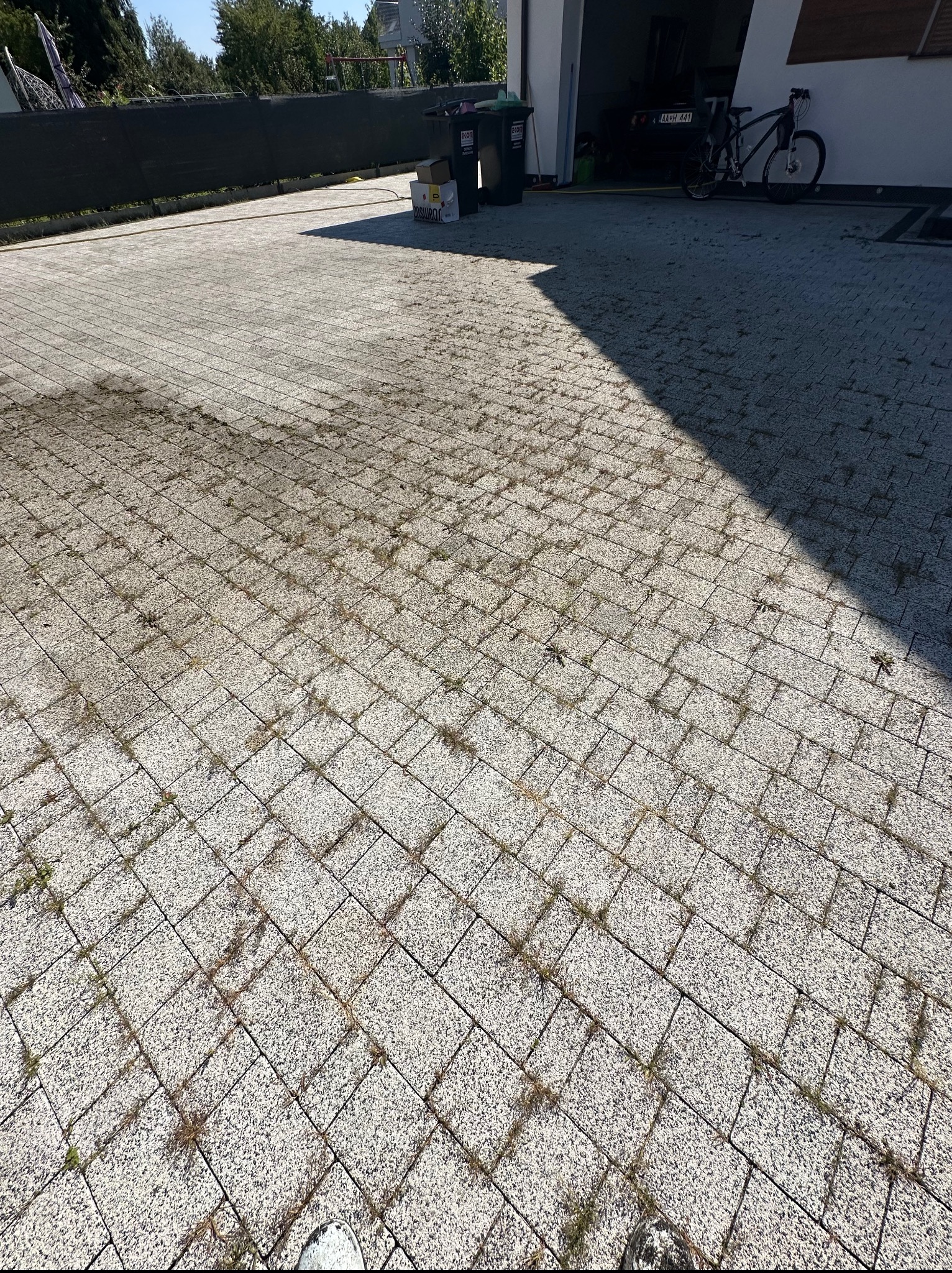 Szara kostka brukowa z chwastami w szparach, częściowo zacieniona, z widocznymi śmietnikami, rowerem i fragmentem budynku w tle. Widok z góry.