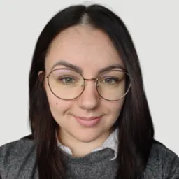 Portret uśmiechniętej brunetki w okularach o złotych oprawkach, ubrana w szary sweter i białą koszulę. Jasne tło, naturalne światło.