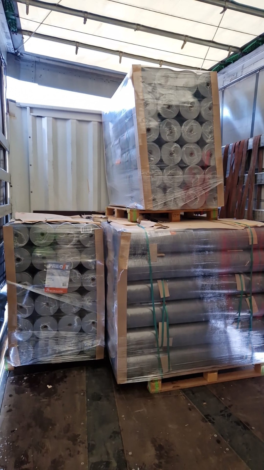 Palety z rolkami materiałów budowlanych zabezpieczone folią i pasami w przestrzeni ładunkowej pojazdu. Widoczne detale opakowania i etykiety na rolkach.