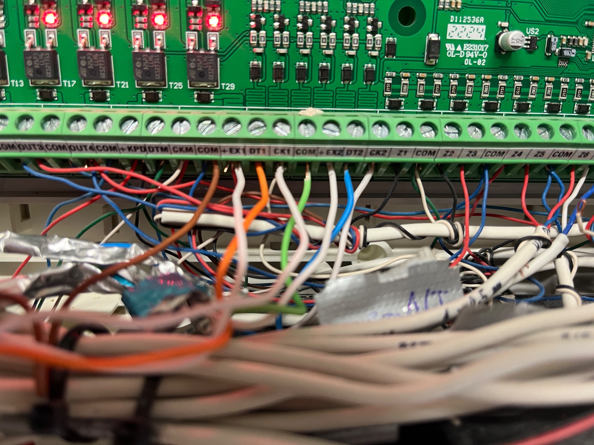 Panel sterowania alarmu z podłączonymi przewodami: czerwonymi, niebieskimi, białymi i pomarańczowymi. Widoczne diody LED i oznaczenia na płycie głównej.