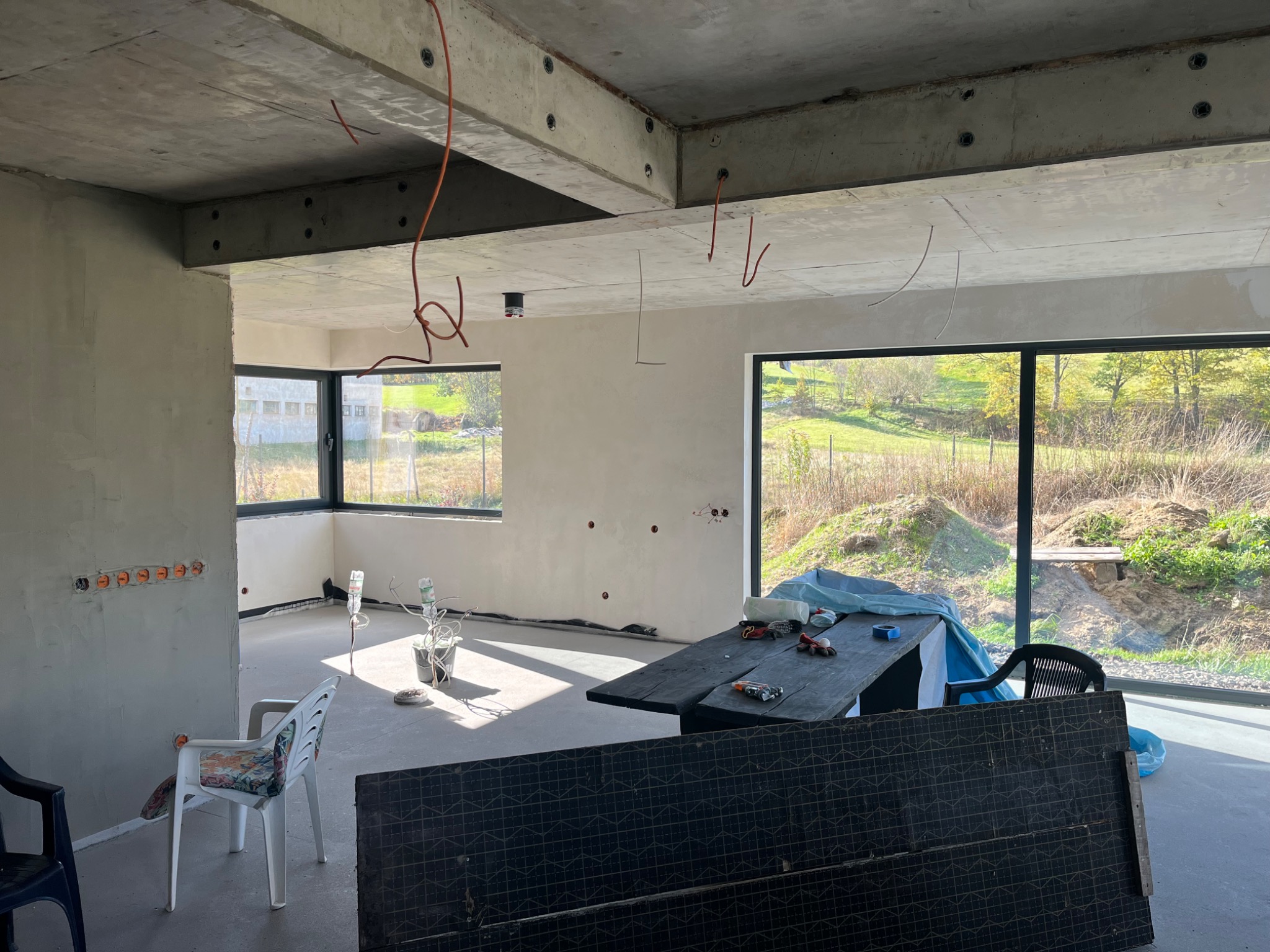 Surowy stan wnętrza z widocznymi przewodami elektrycznymi wystającymi z sufitu i gniazdkami w ścianie. Stolik z narzędziami w pomieszczeniu z dużymi oknami.