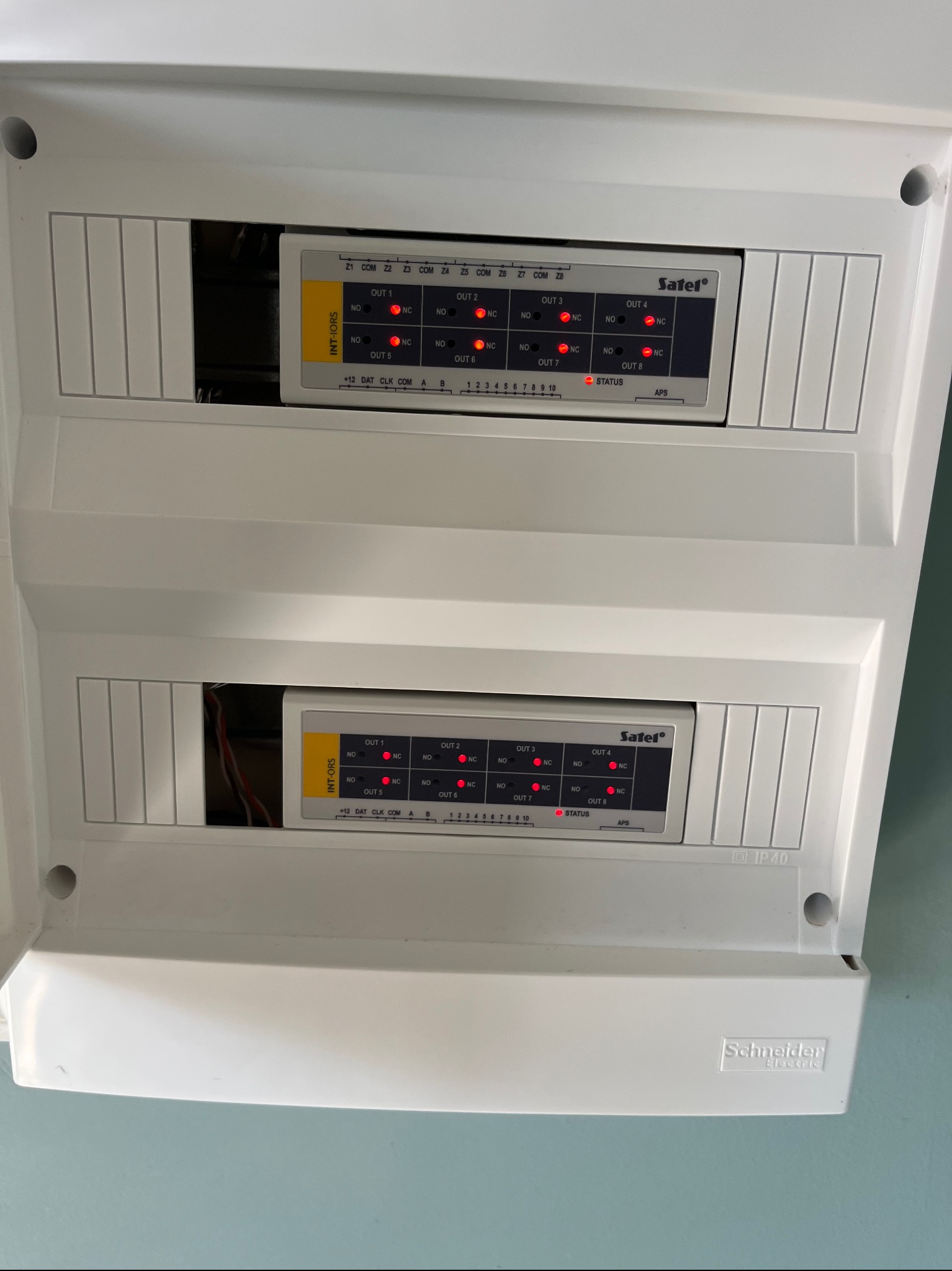 Otwarta, biała skrzynka z dwoma modułami alarmowymi Satel, z diodami LED wskazującymi status. Widoczne kable i oznaczenia na modułach. Schneider Electric IP40.