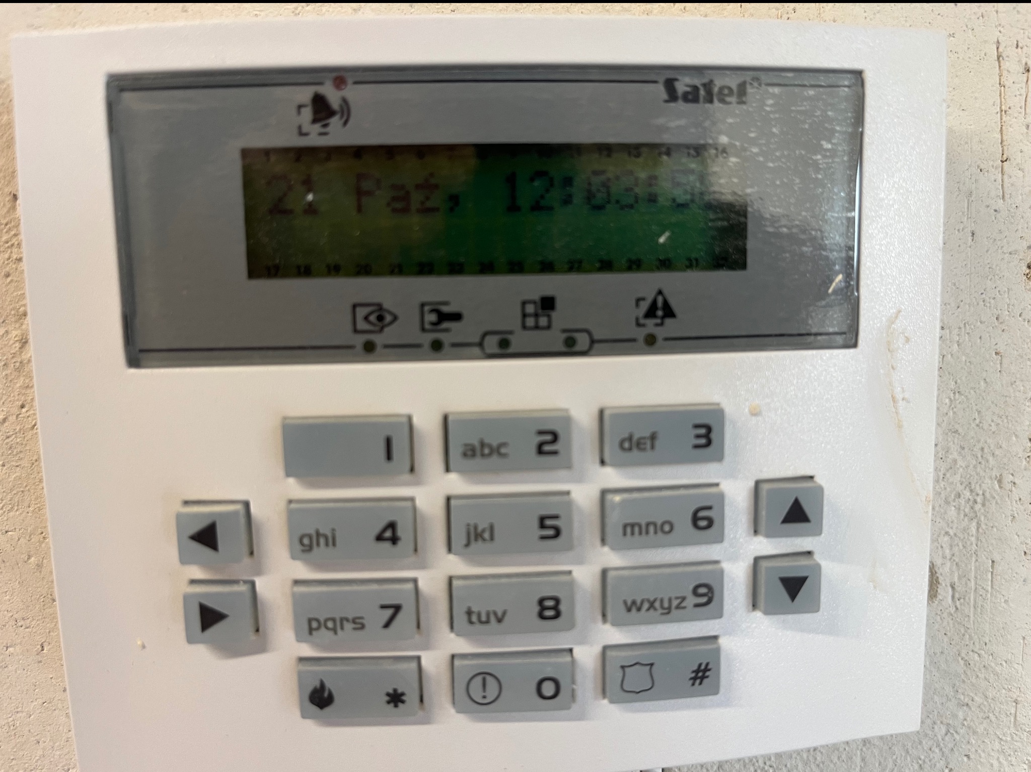 Biały panel sterowania alarmu Satel z wyświetlaczem LCD pokazującym datę i godzinę, umieszczony na szarej ścianie. Widoczne przyciski numeryczne i funkcyjne.