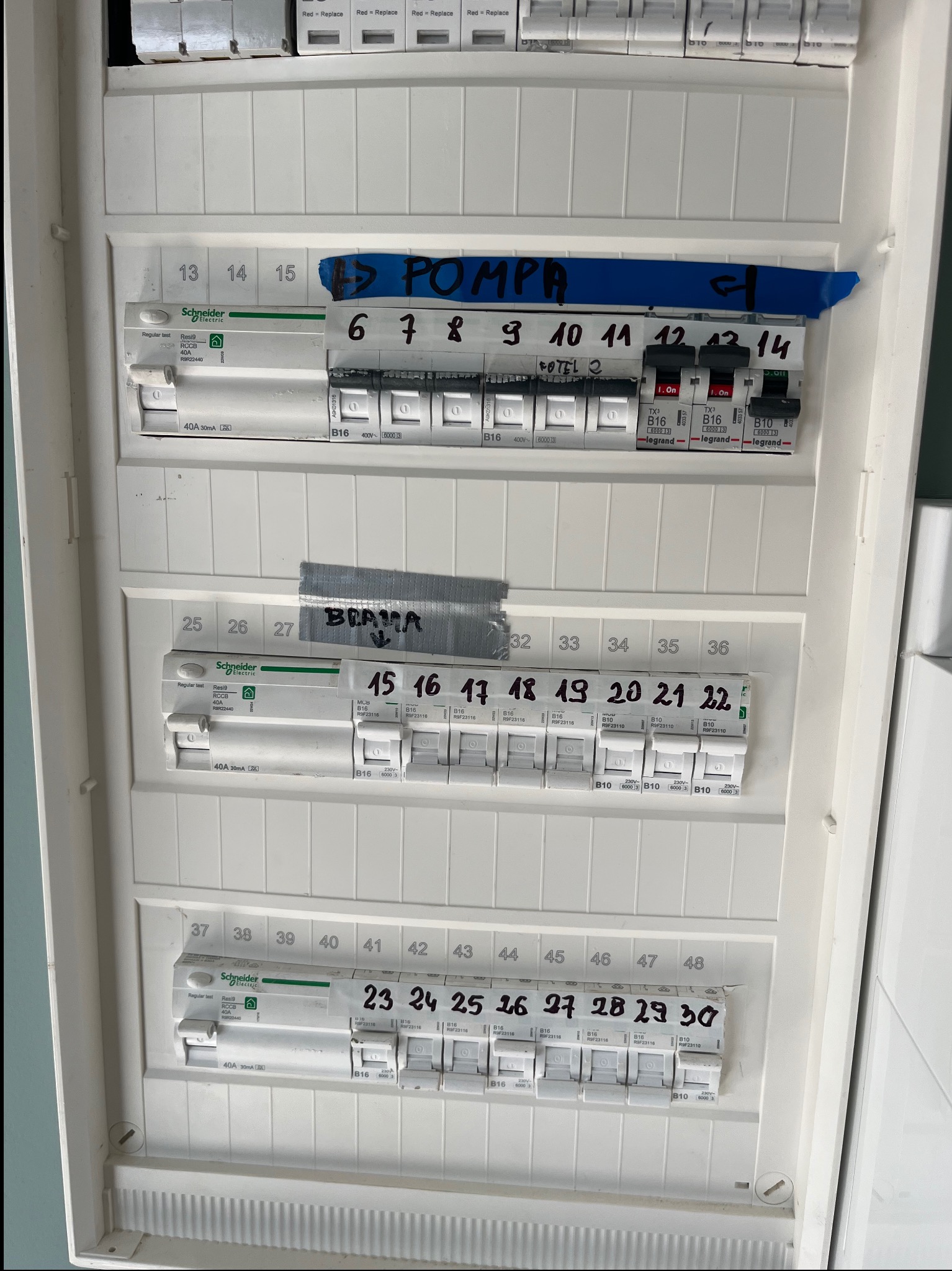 Rozdzielnia elektryczna z bezpiecznikami, opisana taśmą z nazwami obwodów (pompa, brama, fotel). Widoczne oznaczenia i parametry techniczne bezpieczników Schneider Electric i Legrand.