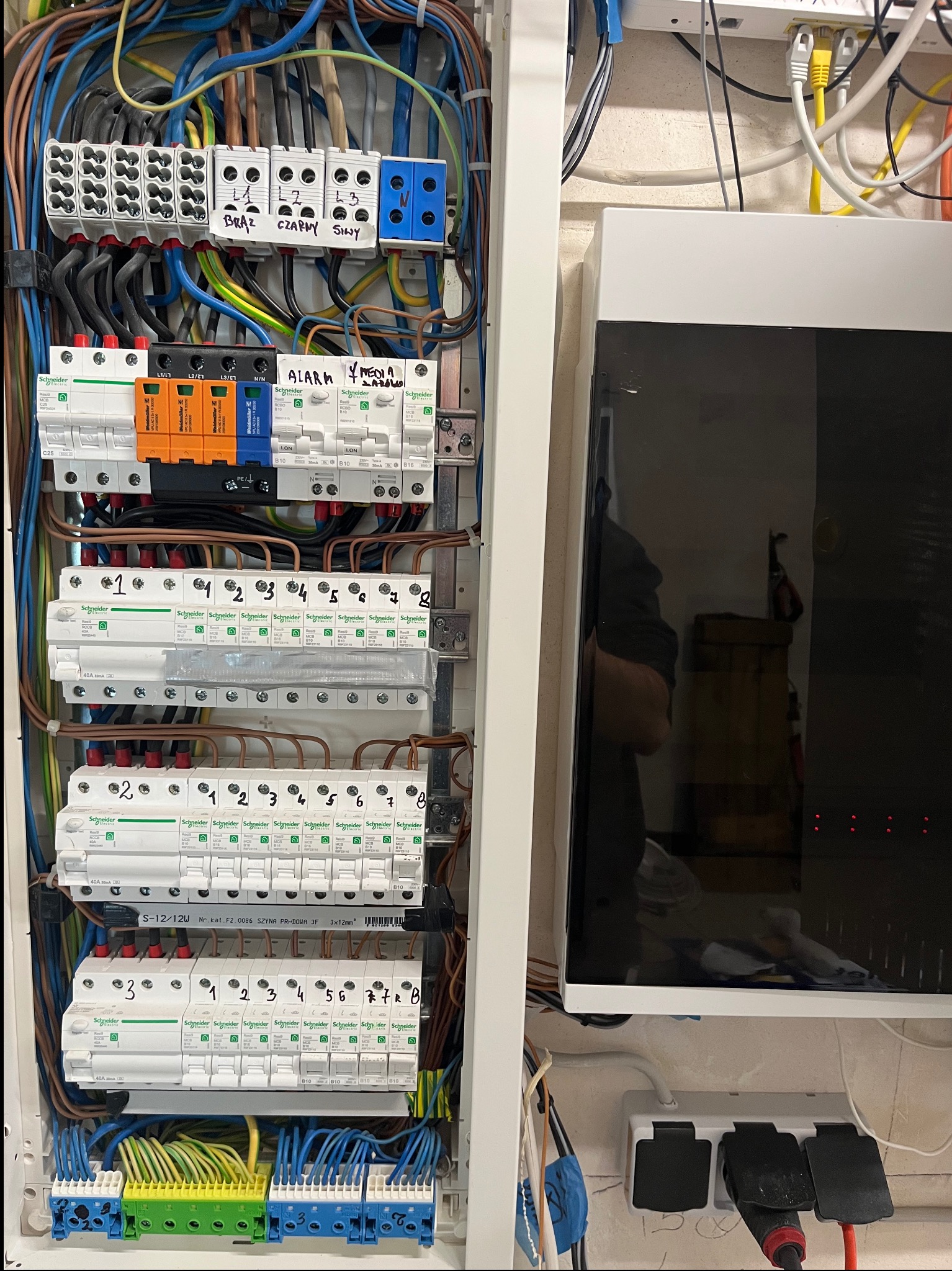 Rozdzielnia elektryczna z bezpiecznikami, okablowaniem i modułem alarmowym. Widoczne oznaczenia kolorów przewodów i numeracja. Obok biała obudowa urządzenia.