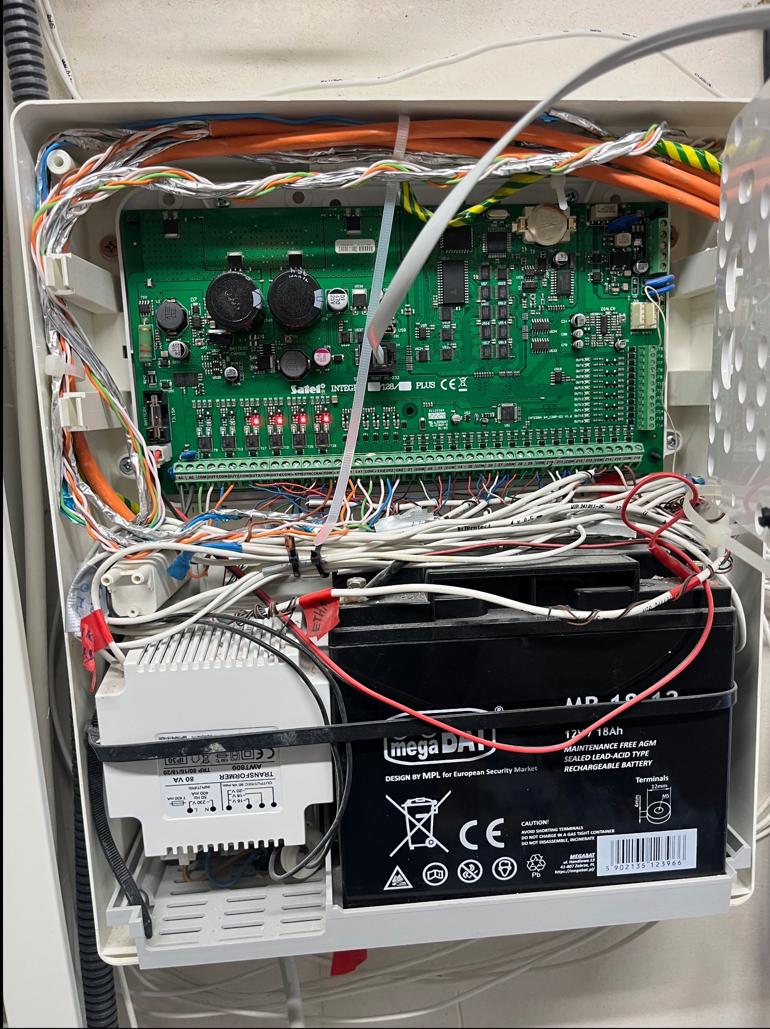Otwarta obudowa centrali alarmowej z widoczną elektroniką, przewodami i akumulatorem. Elementy systemu alarmowego wewnątrz białej skrzynki, widoczne liczne połączenia kablowe.