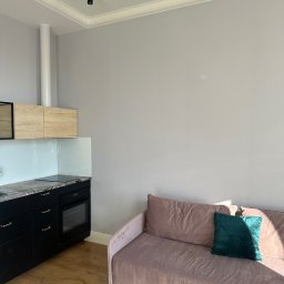 LISSE STUDIO PAWEŁ ŁYSKAWIŃSKI - Jasne wnętrze: czarna zabudowa kuchenna z drewnianymi szafkami wiszącymi, różowa sofa z zieloną poduszką, parkiet i szara ściana. Minimalistyczny design.