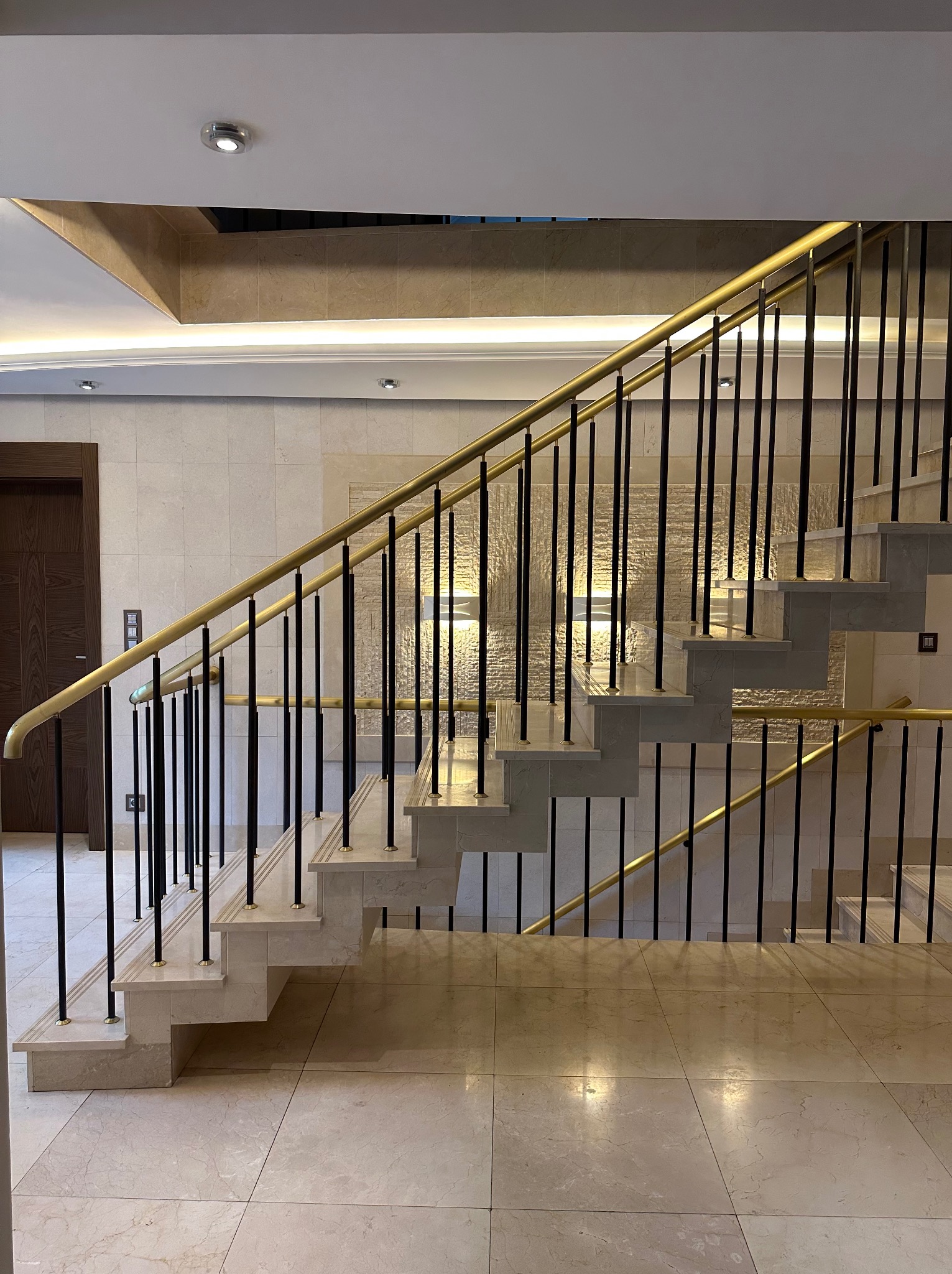 Marmurowe schody z czarnymi balustradami i złotymi poręczami w jasnym wnętrzu. Widoczne płytki podłogowe i drzwi.