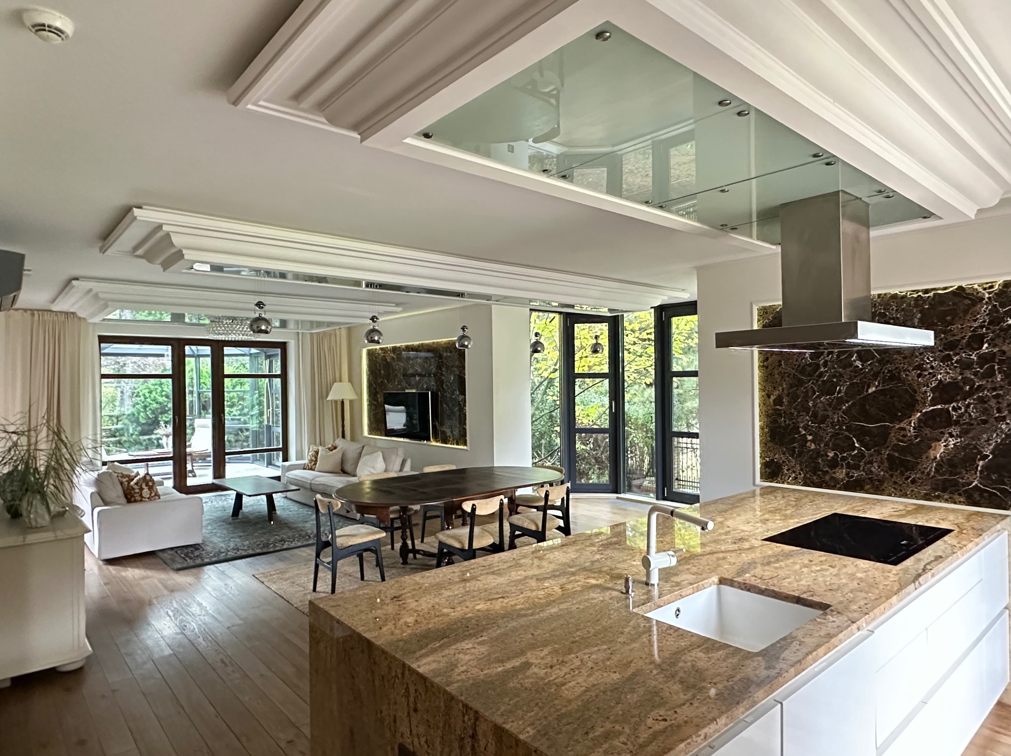 Przestronne wnętrze domu z otwartą kuchnią, jadalnią i salonem. Wnętrze charakteryzuje się eleganckim wykończeniem i dużymi oknami z widokiem na zieleń.