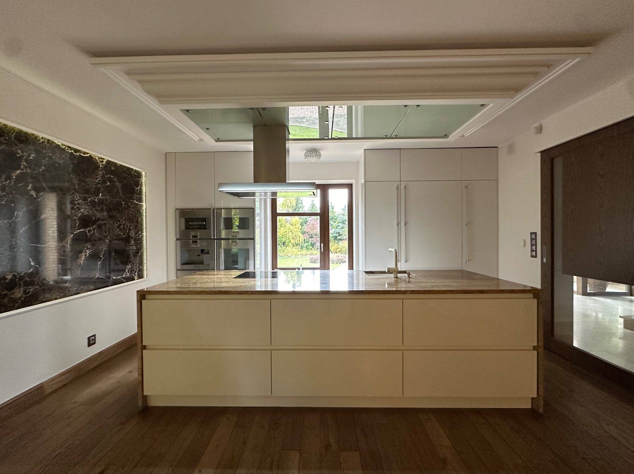 Nowoczesna kuchnia z wyspą i marmurowym akcentem na ścianie. Jasne fronty szafek i drewniana podłoga. Widok na ogród przez przeszklone drzwi.