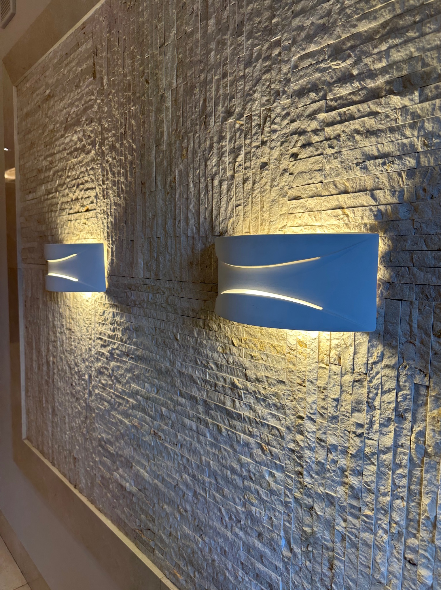 Ściana z kamienia dekoracyjnego oświetlona dwoma nowoczesnymi kinkietami w kształcie prostokątów z zaokrąglonymi bokami, emitującymi ciepłe światło.