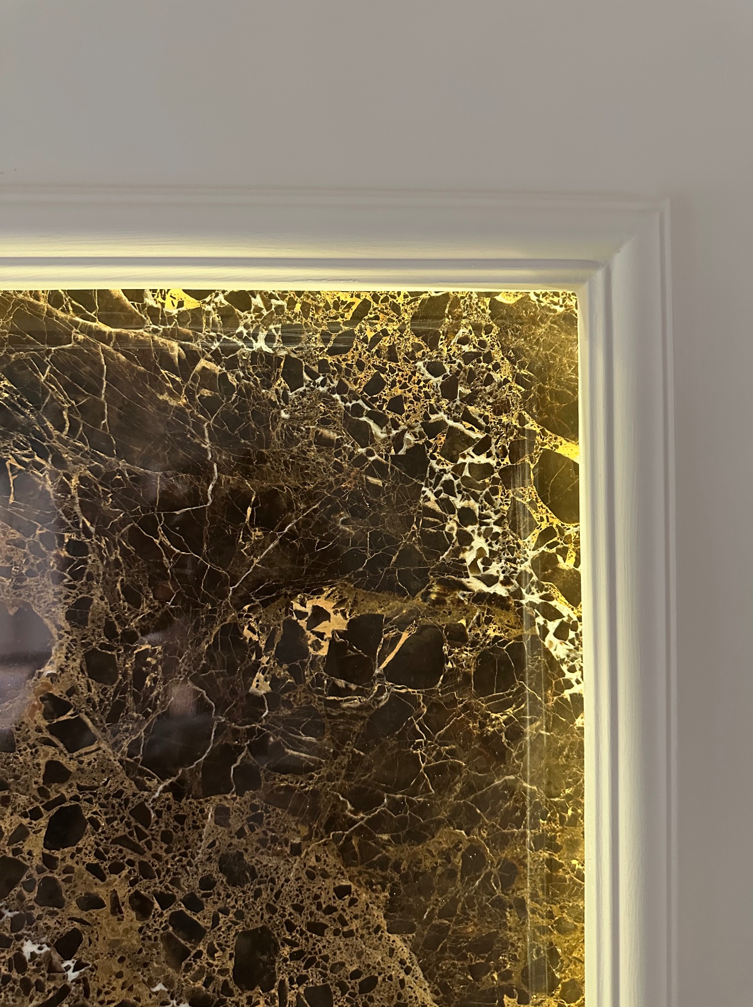 Fragment ściany z dekoracyjnym panelem z czarnego marmuru z złotymi żyłkami, oświetlonym od tyłu, w białej ramie. Detal wykończenia wnętrza.