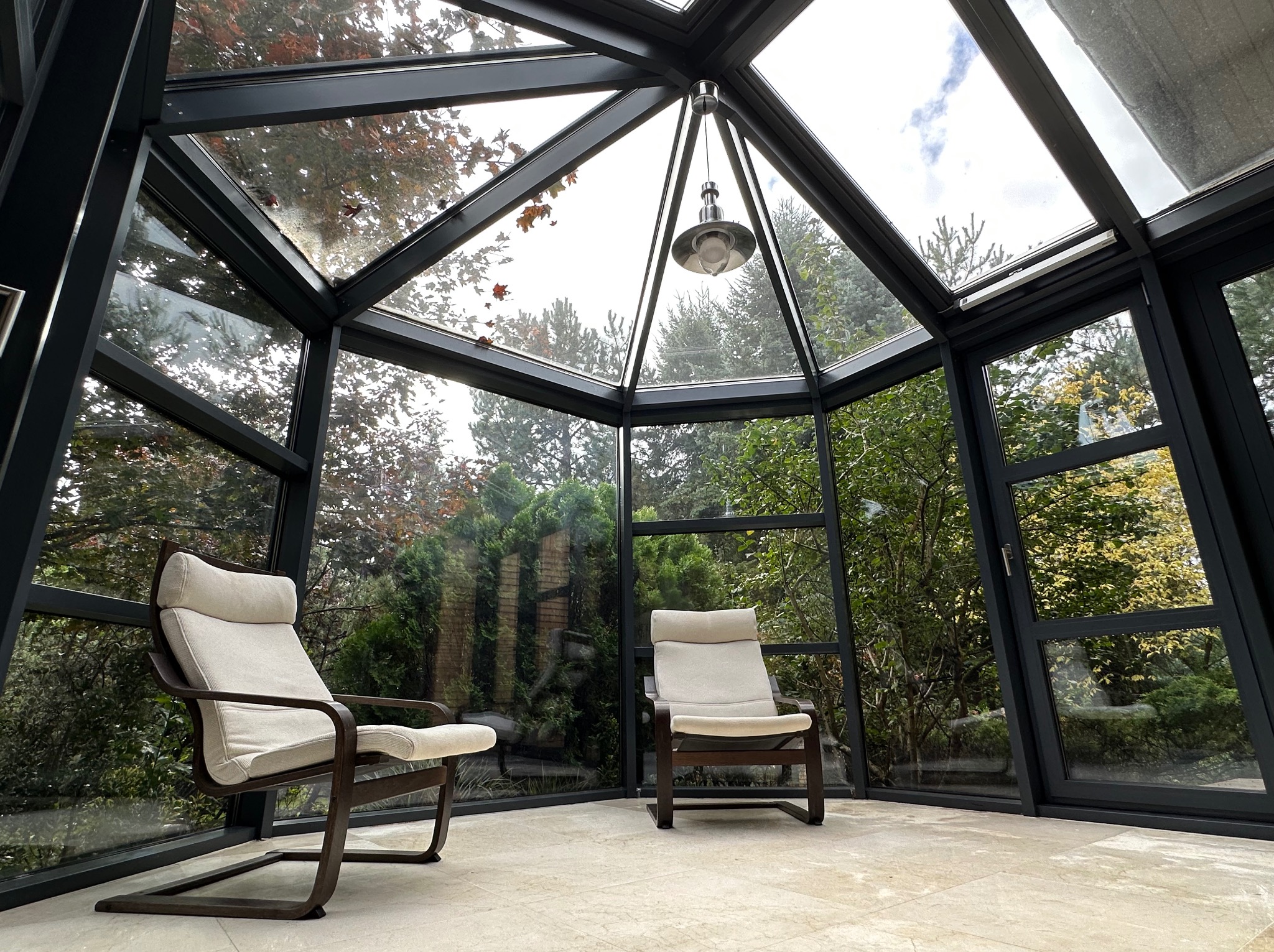 Szklana oranżeria z dwoma fotelami relaksacyjnymi, widok na zieleń ogrodu przez okna. Nowoczesna lampa w centrum pomieszczenia, jasna podłoga.