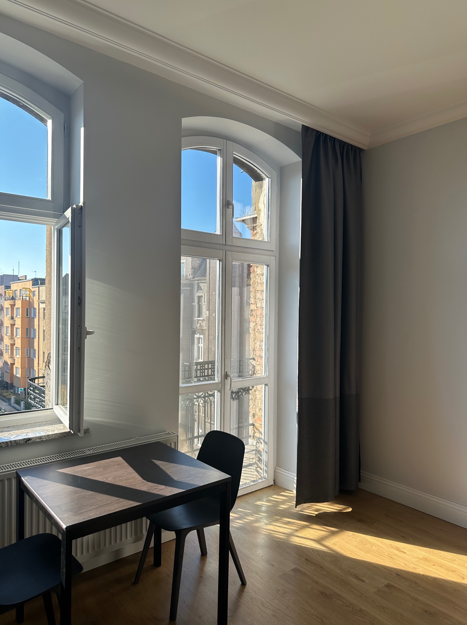 Jasne wnętrze z drewnianą podłogą, stołem i krzesłami. Okna z widokiem na miasto wpuszczają promienie słoneczne. Szare zasłony dodają elegancji.