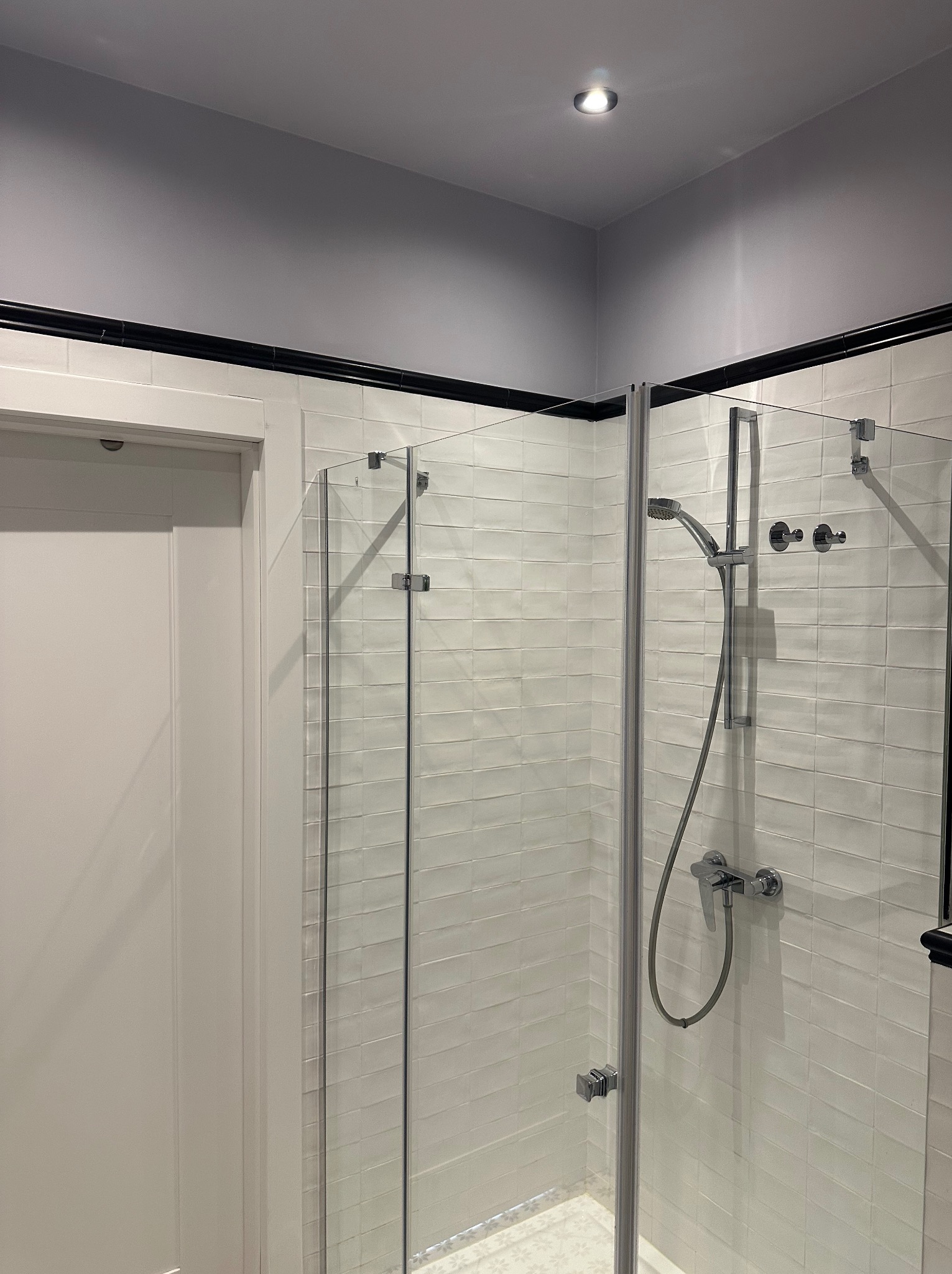 Nowoczesna kabina prysznicowa z białymi, podłużnymi płytkami i czarnymi akcentami. Szklane drzwi z chromowanymi zawiasami. Minimalistyczny design, oświetlenie punktowe.