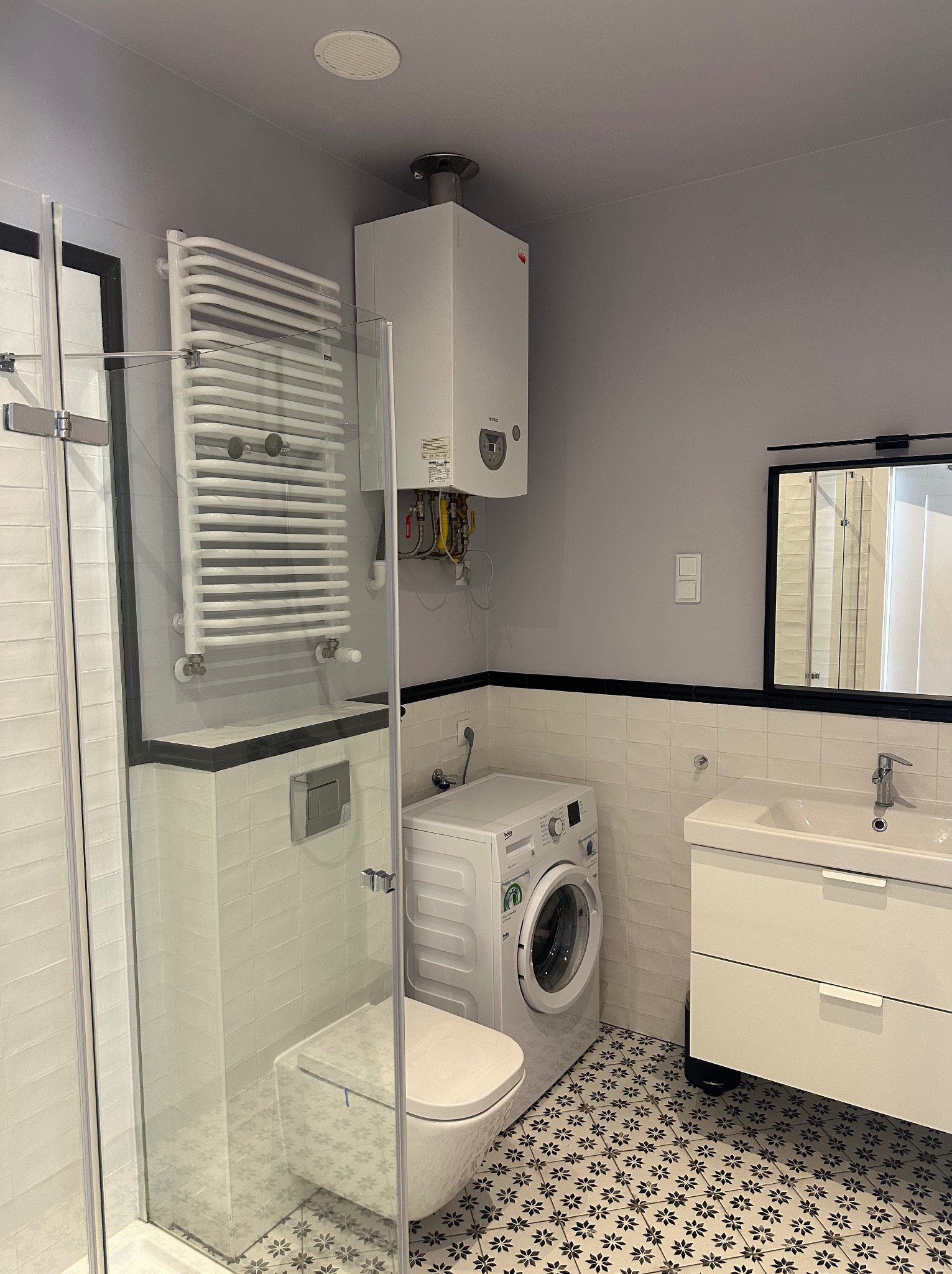 Nowoczesna łazienka po remoncie: białe płytki, wzorzysta podłoga, kabina prysznicowa, umywalka z szafką, pralka i grzejnik drabinkowy. Minimalistyczny design z akcentami czerni.