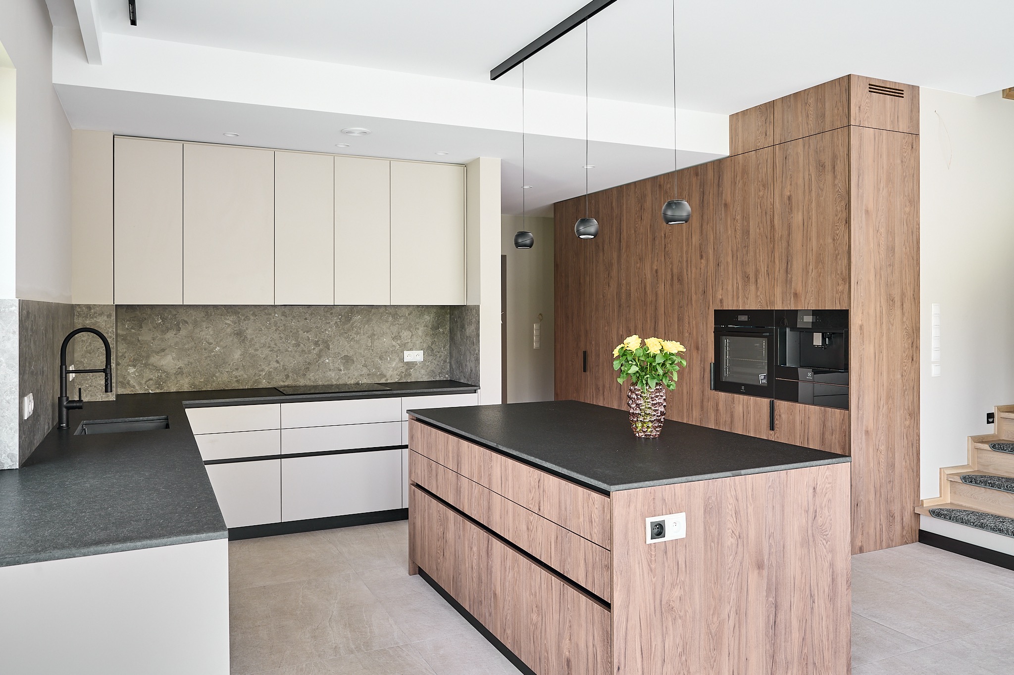 Nowoczesna kuchnia z wyspą i drewnianymi akcentami. Minimalistyczne oświetlenie, szare blaty i marmurowe płytki nadają elegancji. Żółte róże w wazonie ożywiają wnętrze.