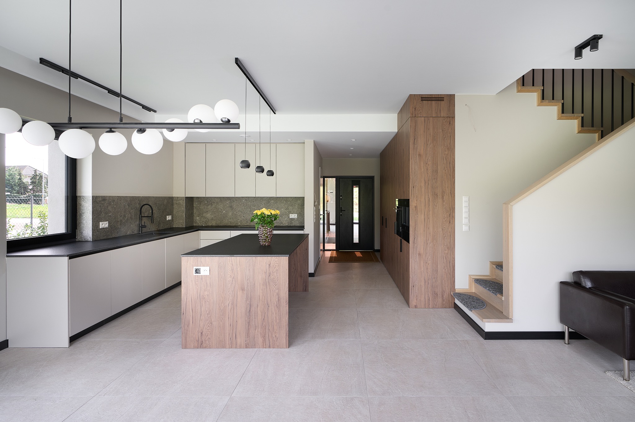 Nowoczesna kuchnia z wyspą i schodami. Jasne szafki, czarny blat, drewniane akcenty. Żółte kwiaty w wazonie na wyspie. Minimalistyczne oświetlenie.