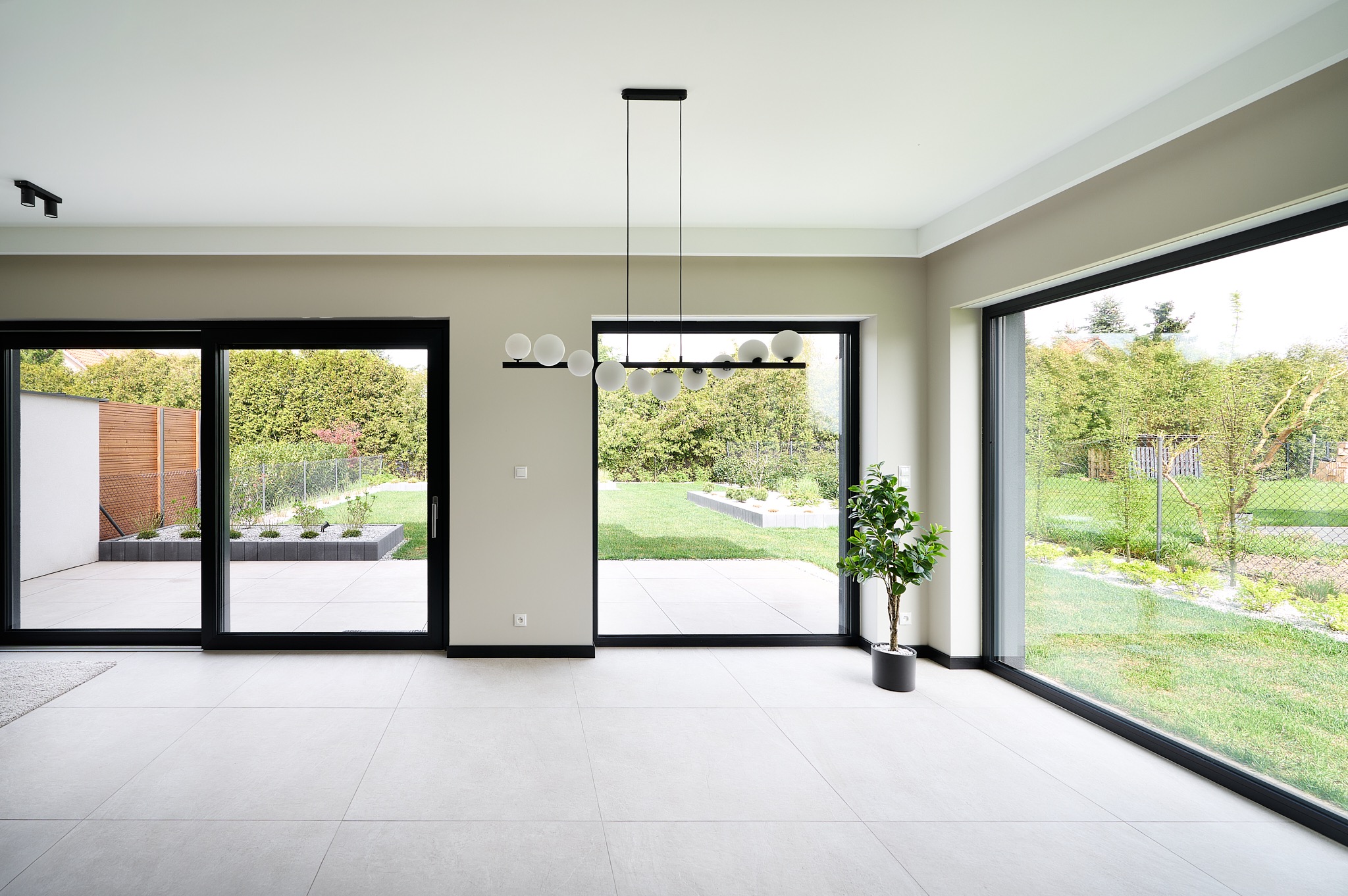 Jasne wnętrze z dużymi oknami i przesuwnymi drzwiami, widok na ogród. Minimalistyczna lampa sufitowa i roślina doniczkowa dodają charakteru. Białe płytki podłogowe.