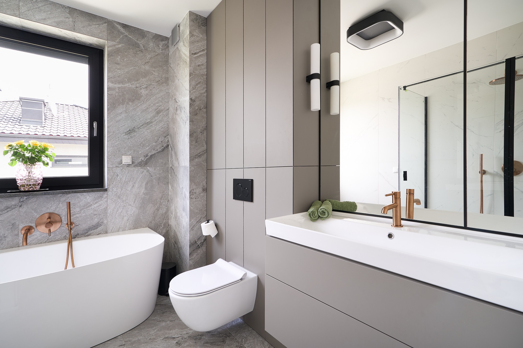 Nowoczesna łazienka z wolnostojącą wanną, szarą armaturą i marmurowymi płytkami. Umywalka z szafką w odcieniach szarości. Okno z widokiem na zewnątrz.