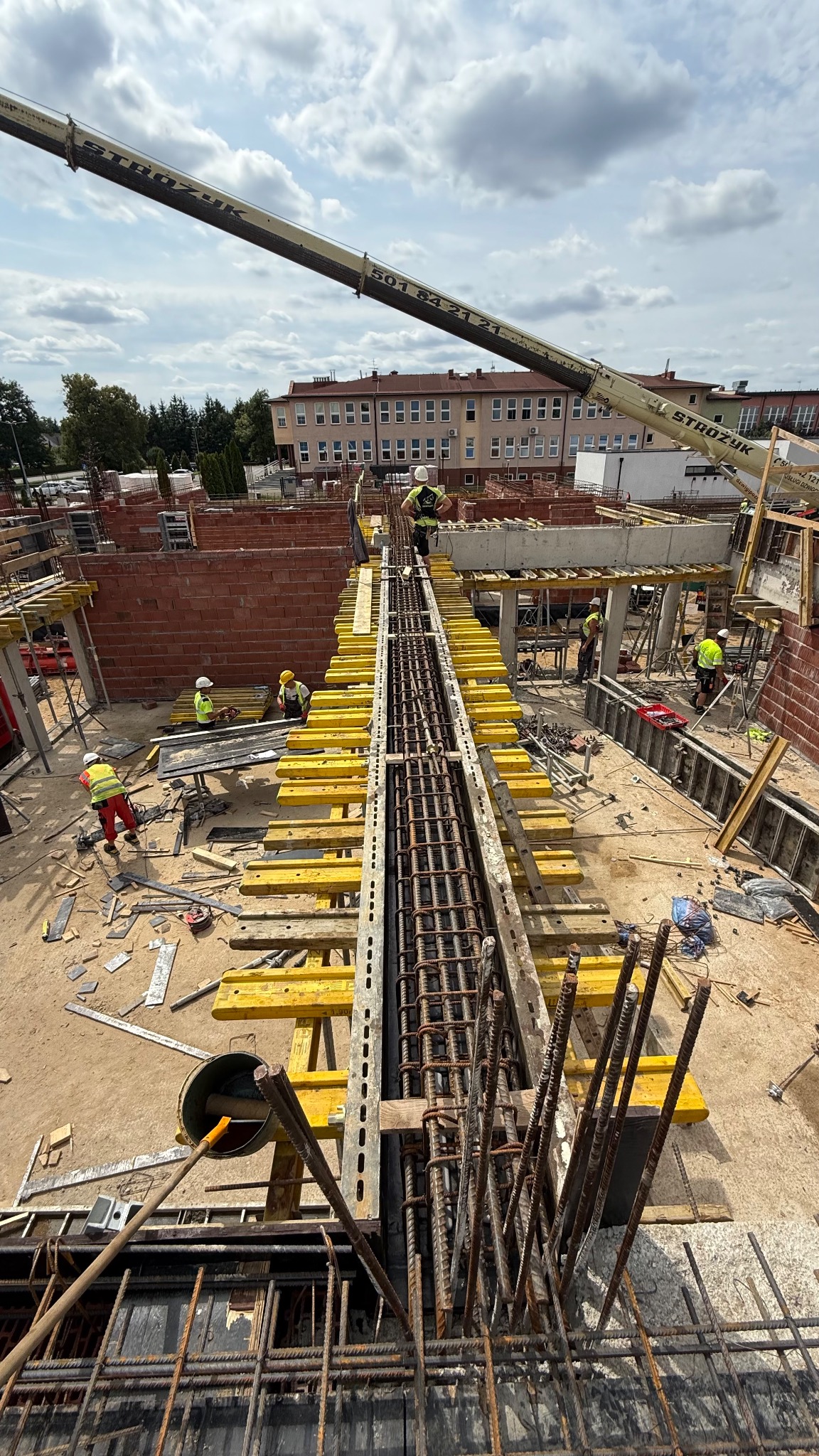 Zbrojenie szkieletu budynku z żółtymi deskami szalunkowymi, widok z góry na plac budowy z ekipą w kaskach i dźwigiem na tle budynku. Etap budowy w słoneczny dzień.