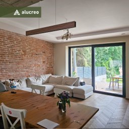ALUCREO SPÓŁKA Z OGRANICZONĄ ODPOWIEDZIALNOŚCIĄ - Salon z ceglaną ścianą i dużym oknem przesuwnym z widokiem na ogród. Nowoczesny design, drewniana podłoga i stół. Pies na kanapie.