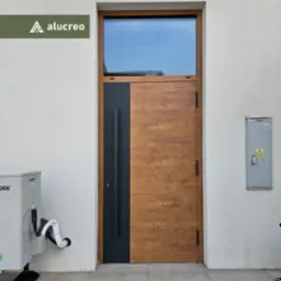 Nowoczesne drzwi wejściowe z drewnianą fakturą i czarnym panelem bocznym, zwieńczone oknem. Widoczna klimatyzacja i skrzynka elektryczna na elewacji.