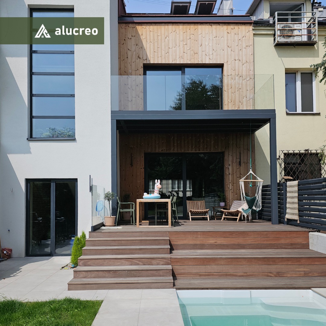 Nowoczesny dom z tarasem i basenem. Widoczne duże przesuwne okna na piętrze i parterze, aluminiowa pergola i szklane balustrady. Drewniane wykończenie elewacji i schody.