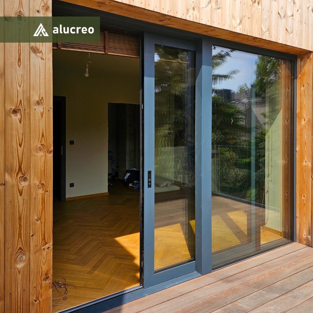 Nowoczesne, szare drzwi tarasowe przesuwne z widokiem na wnętrze i zieleń, zamontowane w drewnianej elewacji, z parkietem w jodełkę wewnątrz.