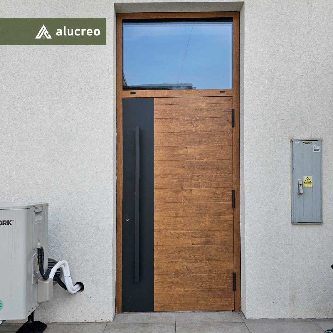 Nowoczesne drzwi wejściowe z drewnianą fakturą i czarnym panelem bocznym, zwieńczone oknem. Widoczna klimatyzacja i skrzynka elektryczna na elewacji.