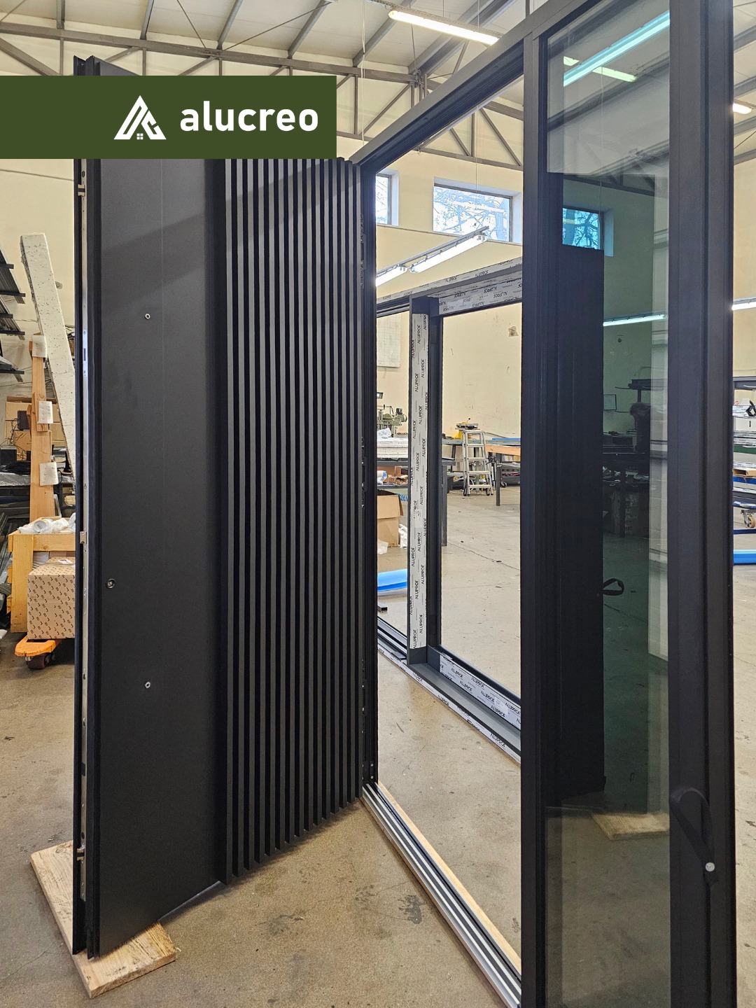 Czarne, częściowo otwarte drzwi przesuwne z pionowymi żłobieniami, widoczne szklane tafle i aluminiowe ramy, w tle hala produkcyjna.