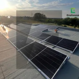 Instalacja fotowoltaiczna na płaskim dachu, widoczne panele w rzędach, w tle budynki mieszkalne i zachodzące słońce. Widoczny cień osoby montującej panele.